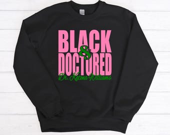 May include: Black and green sweatshirt with the text "Black & Doctored" in pink letters and "Dr. Katerina Williams" in green cursive writing.