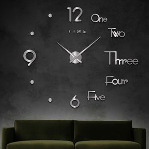 May include: A large, modern wall clock with silver-toned numbers and hands. The clock is mounted on a dark grey wall, and a green sofa is in the foreground. The clock features the words "One", "Two", "Three", "Four", and "Five" instead of the traditional numbers.