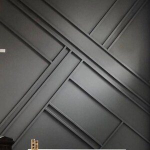 Op de afbeelding: Een donkergrijze muur met een geometrisch patroon van elkaar kruisende lijnen. De muur wordt geaccentueerd door een witte tafel en twee gouden decoratieve objecten.