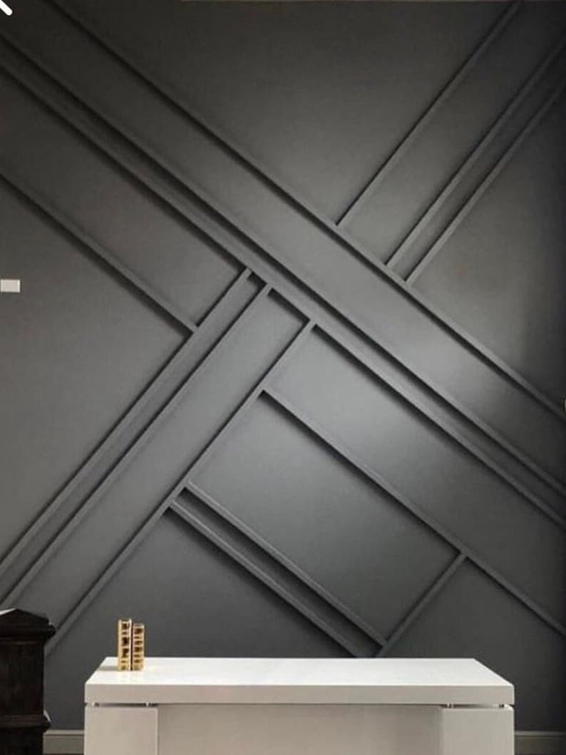 Op de afbeelding: Een donkergrijze muur met een geometrisch patroon van elkaar kruisende lijnen. De muur wordt geaccentueerd door een witte tafel en twee gouden decoratieve objecten.