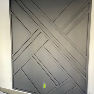 Op de afbeelding: Een donkergrijze muur met een geometrisch patroon gecre&euml;erd door houten stroken. De stroken zijn in een kruispatroon gerangschikt, wat een moderne en stijlvolle uitstraling cre&euml;ert.