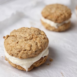 May include: Close-up of two oatmeal cookies with a creamy white filling. The cookies are light brown and have a textured surface. The filling is thick and appears to be frosting or cream. The cookies are on a white surface.