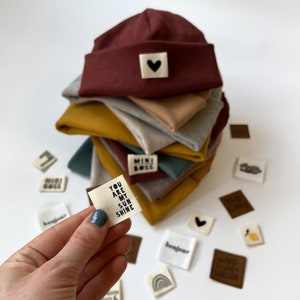 Könnte beinhalten: Ein Stapel gerippter Strickmützen in Kastanienbraun, Grau, Senfgelb und Blaugrün, jeweils mit einem kleinen quadratischen Stoffetikett. Ein im Vordergrund gehaltenes Etikett zeigt den Text YOU ARE MY SUN SHINE, während andere lose Etiketten ein Herz, eine Zitrone, einen Regenbogen und den Text MINI BOSS zeigen.