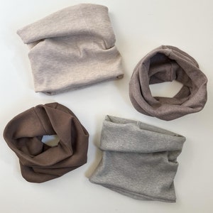 Könnte beinhalten: Vier gerippte Strick-Halswärmer in verschiedenen Farben: Beige, Braun, Grau und Hellgrau. Die Halswärmer sind gefaltet und auf einer weißen Oberfläche angeordnet.