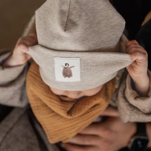 Könnte beinhalten: Eine beige Strickmütze mit einem weißen Aufnäher, auf dem ein Cartoon-Pinguin abgebildet ist. Die Mütze wird von einem Kind gehalten, das einen braunen Pullover und einen braunen und orangen Schal trägt.