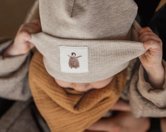 May include: A beige ribbed beanie with a folded brim featuring a square patch with a small brown penguin illustration. Below the beanie is a mustard yellow muslin fabric neck scarf. The beanie has a soft, textured knit construction.