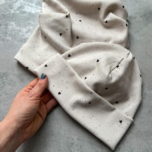 Könnte beinhalten: Zwei beigefarbene Strickmützen mit einem schwarzen Herzmuster. Die Mützen sind gefaltet und übereinander gestapelt.