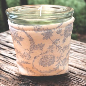 May include: A clear glass jar with a decorative fabric sleeve. The sleeve is a light peach color with a gray floral pattern. The number "1" is written in white on the glass. The jar sits on a wooden surface.