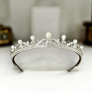 May include: A silver-toned tiara with a curved design, adorned with clear crystals and teardrop-shaped pearls. The tiara features a delicate, ornate pattern and is designed to be worn on the head. The overall theme is bridal or formal wear.