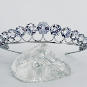 May include: A silver tiara with a row of clear, round, faceted stones. The tiara is on a clear, textured surface.
