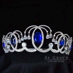 Puede incluir: Una tiara de tono plateado con múltiples piedras azul oscuro de corte ovalado y numerosos cristales transparentes, redondos y en forma de pera, engastados en un marco de metal con volutas. El texto 'de GRAY'S COLLECTION' aparece en blanco abajo a la derecha.
