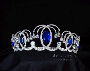 May include: A silver-toned tiara featuring multiple oval-cut deep blue stones and numerous clear, round and pear-shaped crystals set in a swirling metal frame. The text 'de GRAY'S COLLECTION' is printed in white in the lower right corner.