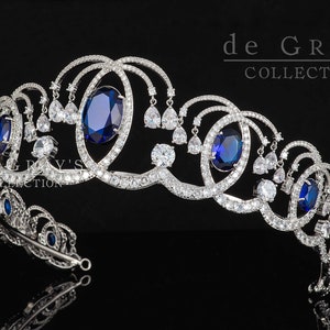 Könnte beinhalten: Eine silberne Tiara mit einem filigranen Design, das mit blauen und klaren Strasssteinen besetzt ist. Die Tiara hat eine geschwungene Form und ist mit mehreren ovalen blauen Steinen verziert. Der Text "de GRAY'S COLLECTION" ist in der oberen rechten Ecke des Bildes sichtbar.