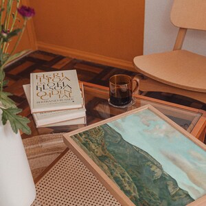 May include: A living room scene with a glass-topped coffee table. On the table are books, a framed landscape painting, and a glass mug with a dark liquid. A white vase with purple flowers and a wooden chair are also visible.