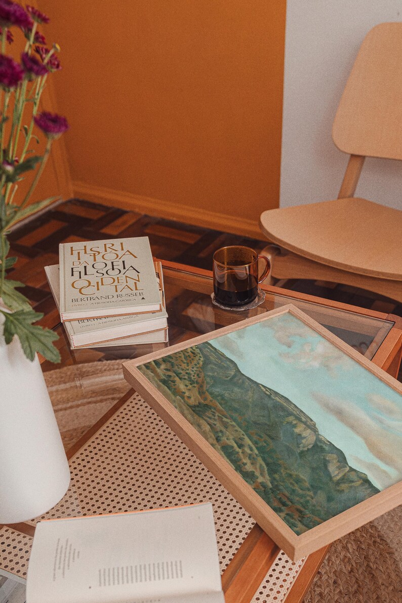 May include: A living room scene with a glass-topped coffee table. On the table are books, a framed landscape painting, and a glass mug with a dark liquid. A white vase with purple flowers and a wooden chair are also visible.