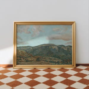 May include: A framed landscape painting with a gold-colored frame. The artwork depicts a mountain range under a cloudy sky. The foreground shows rolling hills with trees. The painting is displayed on a checkered rug.