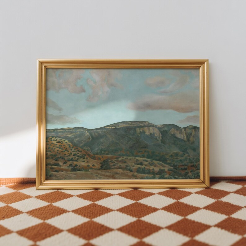 May include: A framed landscape painting with a gold-colored frame. The artwork depicts a mountain range under a cloudy sky. The foreground shows rolling hills with trees. The painting is displayed on a checkered rug.