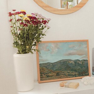 May include: A white vase holds a bouquet of red, yellow, and pink flowers. A framed landscape painting with a wooden frame rests on a white surface. A small paintbrush and decorative items are also present.