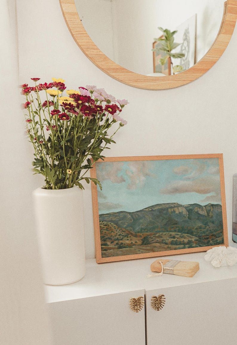 May include: A white vase holds a bouquet of red, yellow, and pink flowers. A framed landscape painting with a wooden frame rests on a white surface. A small paintbrush and decorative items are also present.