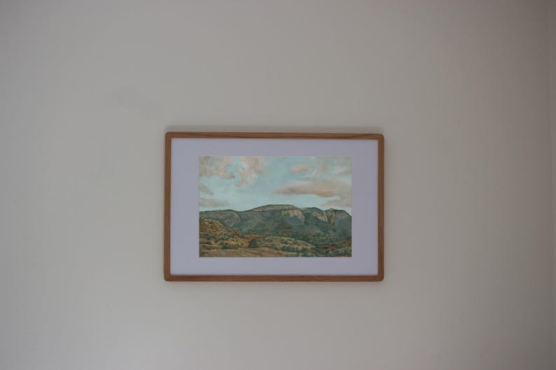 May include: A framed landscape art print featuring a mountain range under a cloudy sky. The artwork has a light brown wooden frame and a white mat. The mountains are green and brown, and the sky has shades of blue and pink.