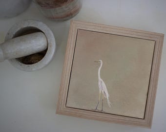 May include: Square framed artwork featuring a white egret on a beige background. The artwork is in a light wood frame. A mortar and pestle and a jar are in the background.