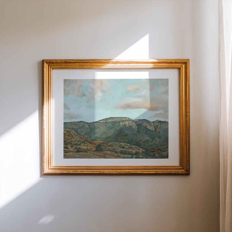 May include: A framed landscape painting featuring a mountain range under a cloudy sky. The artwork is in a gold-colored ornate frame, adding a touch of elegance. The painting depicts a natural scene with green and brown tones.