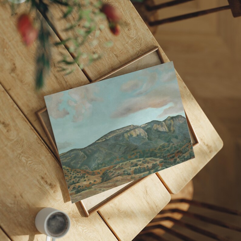 May include: An oil painting of a mountain landscape with a blue sky and pink clouds. The artwork is in a wooden frame and rests on a wooden table. A white mug is visible in the lower left corner.