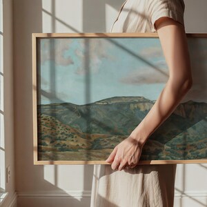 May include: A framed landscape painting depicting a mountain range under a cloudy sky. The artwork features a natural color palette of greens, browns, and blues. The frame is made of light-colored wood.