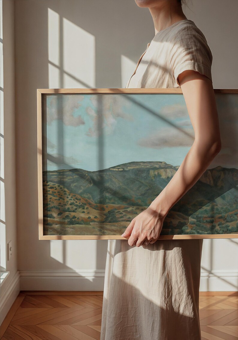 May include: A framed landscape painting depicting a mountain range under a cloudy sky. The artwork features a natural color palette of greens, browns, and blues. The frame is made of light-colored wood.