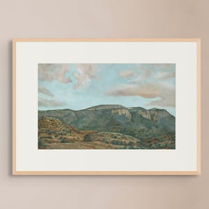 May include: A framed landscape painting depicts a mountain range under a blue sky with fluffy clouds. The artwork features a palette of greens, browns, and blues, creating a serene and naturalistic scene. The frame is a light wood color.
