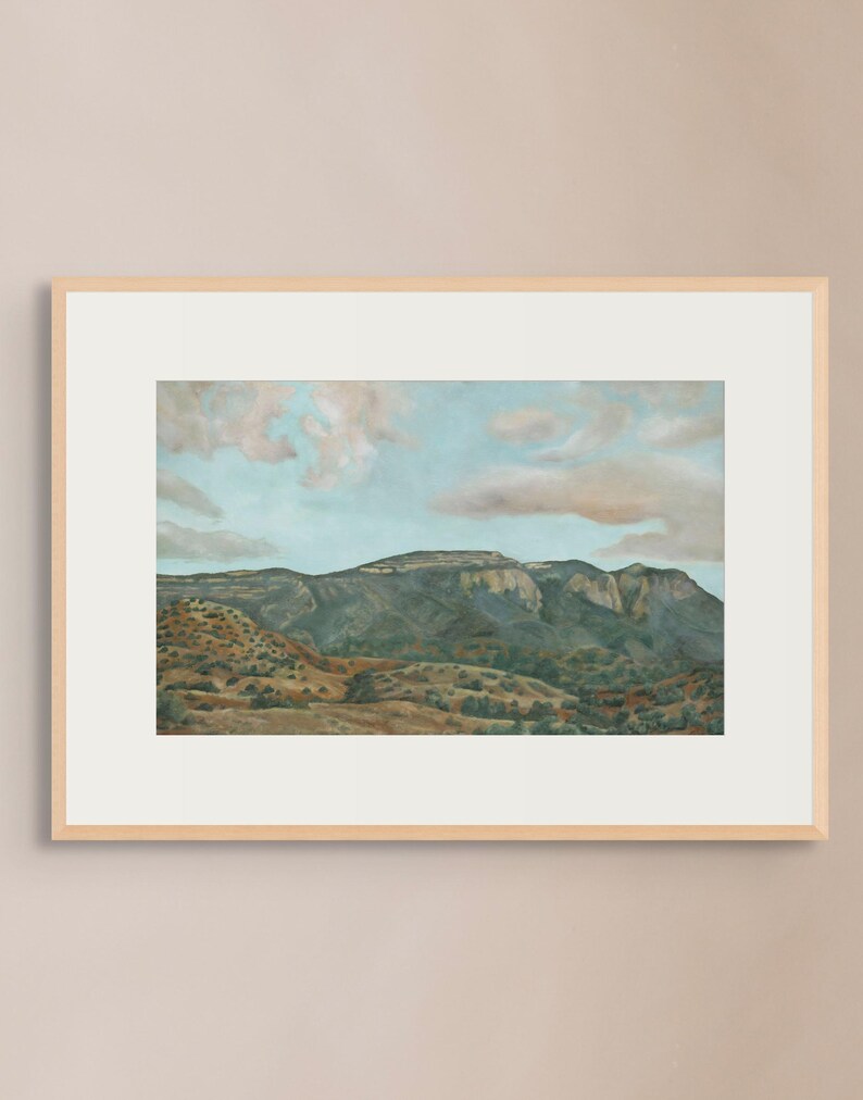May include: A framed landscape painting depicts a mountain range under a blue sky with fluffy clouds. The artwork features a palette of greens, browns, and blues, creating a serene and naturalistic scene. The frame is a light wood color.