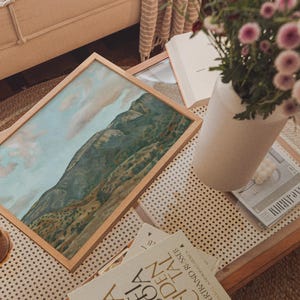 May include: A framed landscape painting of a mountain range with a light wood frame rests on a glass-topped coffee table. A vase of pink flowers, books, and a glass of tea are also on the table, creating a cozy, artistic scene.