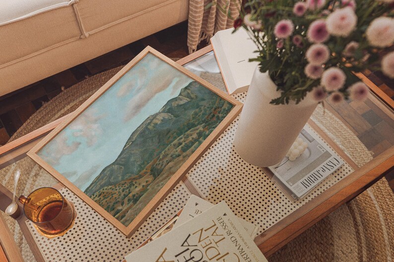 May include: A framed landscape painting of a mountain range with a light wood frame rests on a glass-topped coffee table. A vase of pink flowers, books, and a glass of tea are also on the table, creating a cozy, artistic scene.