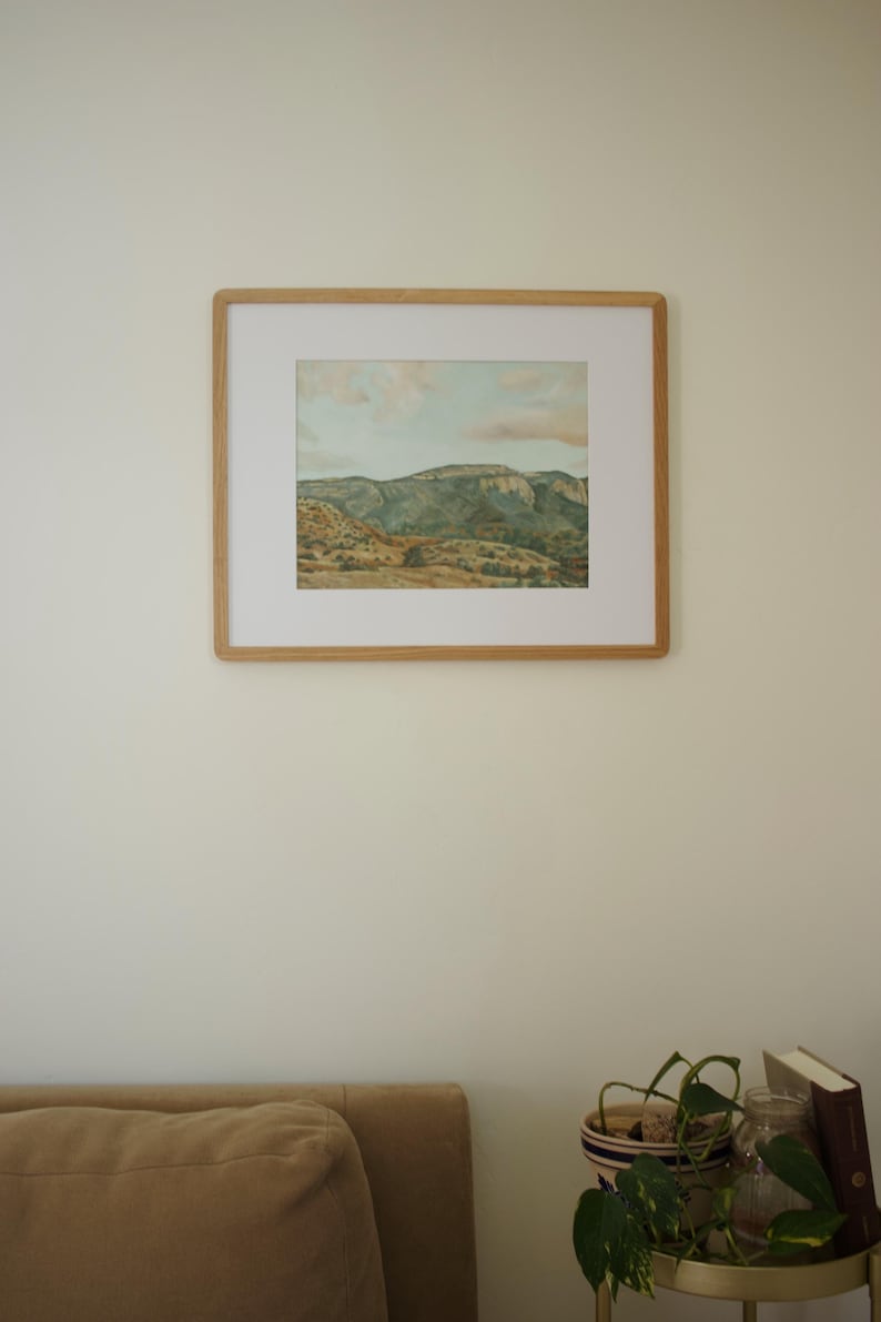 May include: A framed landscape art print depicting a mountain range under a cloudy sky. The artwork is in a light wood frame and hangs on a cream-colored wall. The color palette includes greens, browns, and blues.