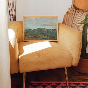 May include: A framed landscape painting of a mountain range with a blue sky and clouds. The artwork is displayed on a mustard-yellow velvet armchair with gold legs. The scene is illuminated by natural light, creating shadows.