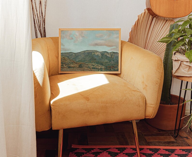 May include: A framed landscape painting of a mountain range with a blue sky and clouds. The artwork is displayed on a mustard-yellow velvet armchair with gold legs. The scene is illuminated by natural light, creating shadows.