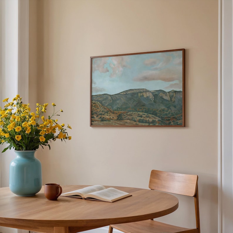 May include: A dining room scene featuring a round wooden table, a wooden chair, and a framed landscape painting. A light blue vase holds yellow flowers. A brown mug and an open book are on the table. The painting depicts a mountain range under a cloudy sky.