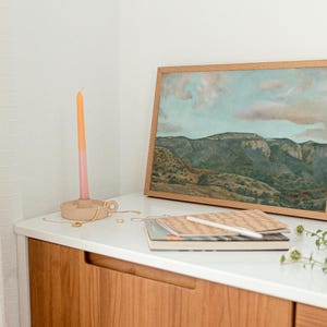 May include: A framed landscape painting with a mountain range and cloudy sky sits on a white surface. A wooden candle holder with a pink and orange candle, notebooks, and a vase with dried stems are also on the surface.