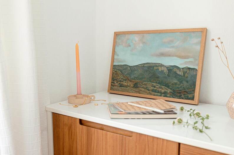 May include: A framed landscape painting with a mountain range and cloudy sky sits on a white surface. A wooden candle holder with a pink and orange candle, notebooks, and a vase with dried stems are also on the surface.