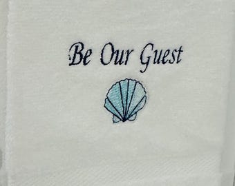 以下が含まれることがあります： 白いバスタオル。筆記体で「Be Our Guest」の刺繍が施され、水色の貝殻のデザインが施されています。タオルはバスルームの銀色のバーに掛けられています。