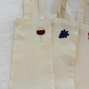 以下が含まれることがあります： クリーム色のキャンバスワインボトルバッグ、刺繍デザイン入り。バッグには、ボルドー色のワインが入ったワイングラス、青いブドウの房、赤いイチゴが描かれています。各バッグには、お揃いのクリーム色のハンドルが付いています。