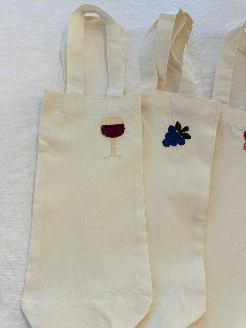 以下が含まれることがあります： クリーム色のキャンバスワインボトルバッグ、刺繍デザイン入り。バッグには、ボルドー色のワインが入ったワイングラス、青いブドウの房、赤いイチゴが描かれています。各バッグには、お揃いのクリーム色のハンドルが付いています。