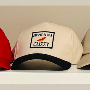 May include: A cream-colored baseball cap with a black brim. The cap features a rectangular patch with the text "BAD DAY TO BE A GLIZZY" and a graphic of a hot dog.