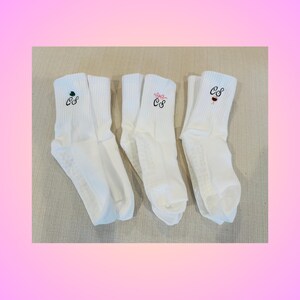 May include: Three pairs of white athletic socks with non-slip soles. Each sock features embroidered initials "CS" with different embellishments: a green shamrock, a pink bow, and a red lipstick.