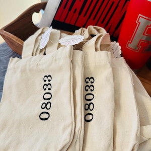 May include: Several small, off-white canvas tote bags with black embroidered numbers "08033". The bags have short handles and are stacked together. A red tumbler with a silver "H" logo and a black and red design are in the background.