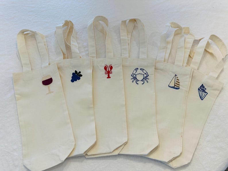 以下が含まれることがあります： オフホワイトのキャンバスワインボトルバッグのコレクション。各バッグには、ワイングラス、ブドウ、ロブスター、カニ、ヨット、貝殻など、異なる刺繍デザインが施されています。バッグにはお揃いのハンドルが付いています。