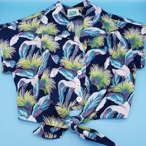 May include: A navy blue short-sleeved shirt with a tropical leaf print. The shirt has a tie front and three buttons. The shirt is made of a lightweight fabric and is perfect for warm weather.