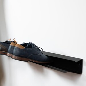 Peut inclure: Un porte-chaussures en m&eacute;tal noir mont&eacute; sur un mur blanc avec deux paires de chaussures dessus. Les chaussures sont une paire en cuir bleu fonc&eacute; et une paire en cuir marron.