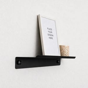 Könnte beinhalten: Ein schwarzes Metall-Wandregal mit rechteckigem Design. Ein weiß gerahmter Druck mit dem Text "PLACE YOUR DESIGN HERE" lehnt an der Wand. Ein kleiner, dekorativer Kerzenhalter steht auf dem Regal.