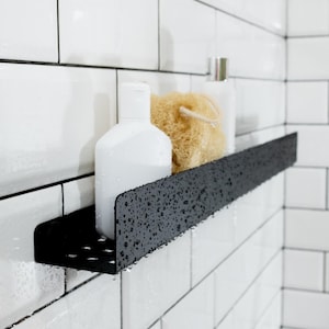 May include: A black metal shower shelf with a textured finish. The shelf is mounted on a white tiled wall and holds two bottles of shampoo and a loofah.
